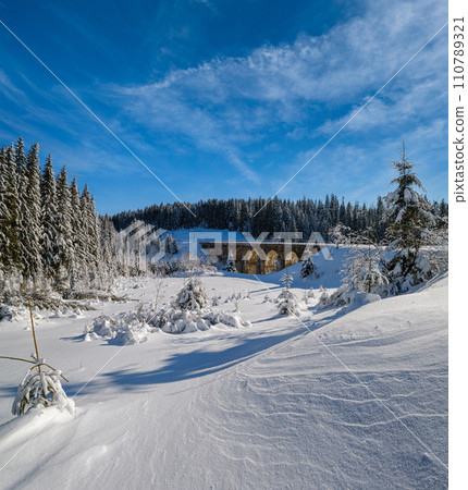 Stone viaduct (arch bridge) on railway through mountain snowy fir forest. Snow drifts  on wayside and hoarfrost on trees and electric line wires. 110789321