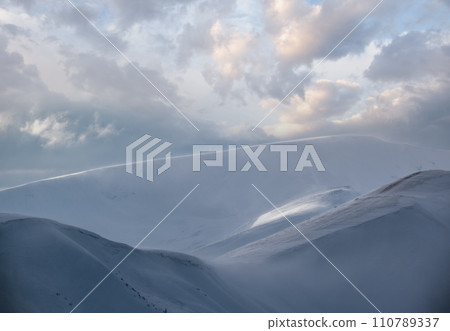 Snow covered winter mountains  in last evening sunlight. Magnificent windy dusk on tops above picturesque alpine ski resort 110789337