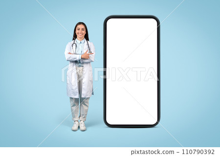 Young female doctor in white coat standing beside huge cellphone screen, pointing at blank screen, medical info display Young female doctor in white coat standing beside huge cellphone screen, pointing at blank screen, medical info display 110790392