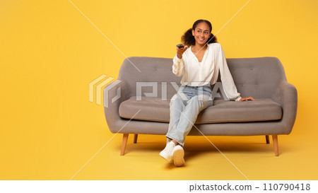 Black teenager girl watching TV sitting on couch, yellow backdrop 110790418