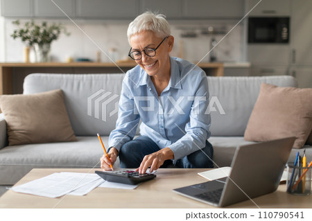 Joyful pensioner mature lady at home calculating budget on calculator Joyful pensioner mature lady at home calculating budget on calculator 110790541
