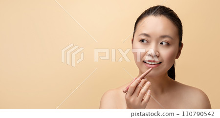 Cheerful millennial chinese naked woman applies pencil to lips and makes basic nude makeup 110790542