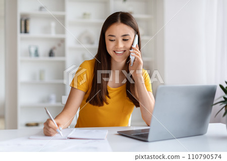 Home Office. Young Asian Woman Talking On Cellphone And Taking Notes Home Office. Young Asian Woman Talking On Cellphone And Taking Notes 110790574