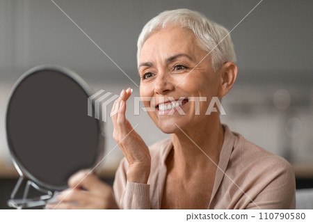 Cheerful Senior Lady Applying Facial Moisturizer Looking At Mirror Indoor Cheerful Senior Lady Applying Facial Moisturizer Looking At Mirror Indoor 110790580