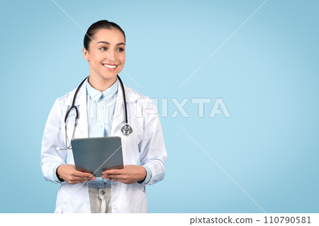 Cheerful young female doctor in white coat holding digital tablet, looking at copy space over blue studio background Cheerful young female doctor in white coat holding digital tablet, looking at copy space over blue studio background 110790581