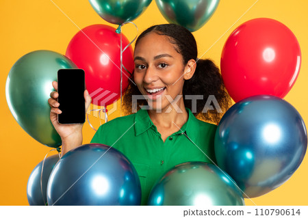 Happy black teenager girl poses with cellphone among balloons, studio 110790614