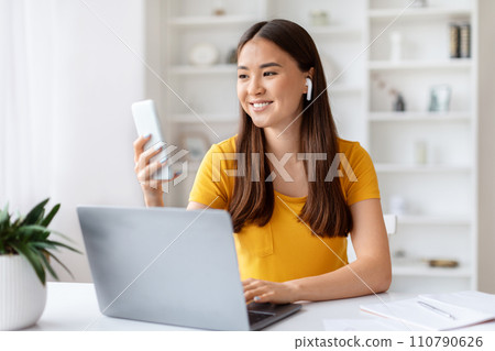 Smiling Asian woman multitasking with smartphone and laptop in home office Smiling Asian woman multitasking with smartphone and laptop in home office 110790626
