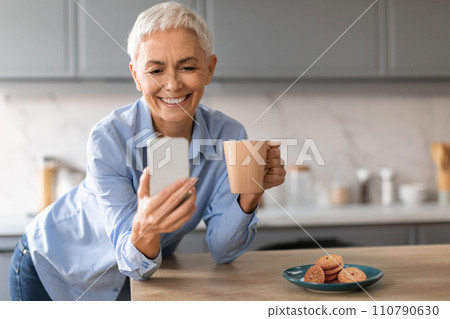 Relaxed mature lady holding smartphone and cup of coffee indoor 110790630