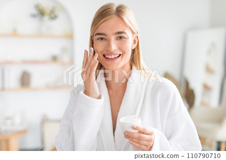 blonde woman smiling to camera while applies moisturizer in bathroom 110790710