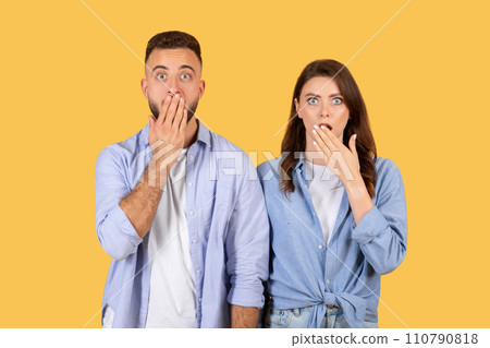 Shocked couple covering mouths, wide-eyed surprise on yellow background 110790818