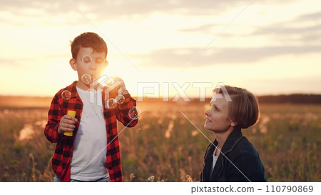 Son blows soap bubbles standing in field with growing plants near mother 110790869