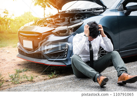 Young businessman wearing white shirt and call roadside assistance. Worried mature man holding his head by hands sitting near his broken car with raised hood on the roadside of a rural road. Young businessman wearing white shirt and call roadside assistance. Worried mature man holding his head by hands sitting near his broken car with raised hood on the roadside of a rural road. 110791065