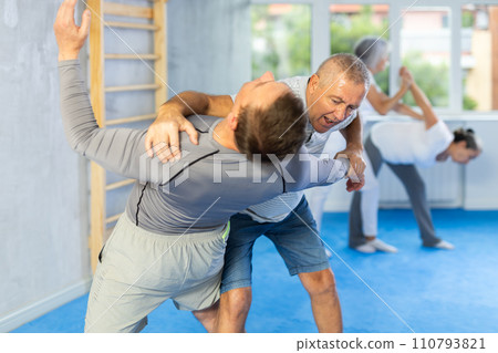 Mature man at self-defense lesson learns how to make painful blows with his hand and foot Mature man at self-defense lesson learns how to make painful blows with his hand and foot 110793821