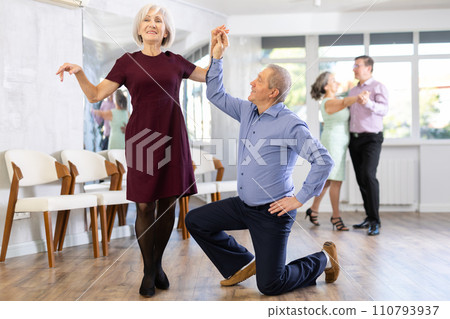 Happy mature couple enjoying retro ballroom dancing in dance salon, man standing on knee and holding hand of woman Happy mature couple enjoying retro ballroom dancing in dance salon, man standing on knee and holding hand of woman 110793937