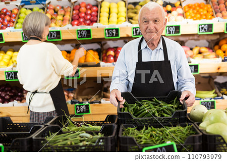 Elderly supermarket employee rearranges box of green chili peppers 110793979