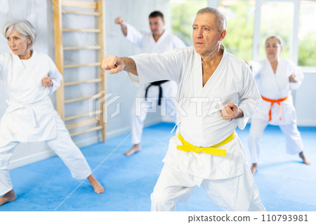 Elderly women and men in kimonos stand in a fighting stance during group karate training session 110793981