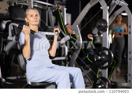 Focused elderly woman exercising on shoulders press machine 110794003