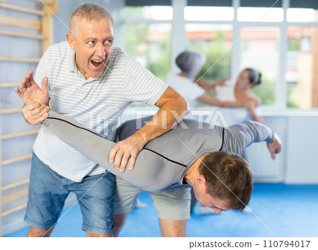 Self defense lesson - elderly man twists the arm of attacking man with painful hold in gym 110794017