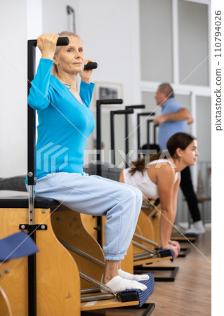 Concentrated mature female practicing Pilates system on Wanda chair machine in studio to improve mobility of upper back Concentrated mature female practicing Pilates system on Wanda chair machine in studio to improve mobility of upper back 110794026