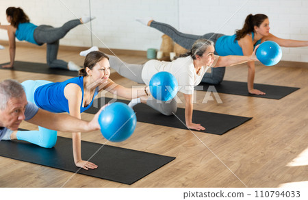 People of different ages stretching leg and arm, holding mini ball in their hands during Pilates workout in studio People of different ages stretching leg and arm, holding mini ball in their hands during Pilates workout in studio 110794033