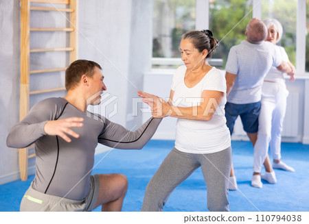 Elderly woman twists arm of attacking man with painful hold in gym. Self-defense lesson Elderly woman twists arm of attacking man with painful hold in gym. Self-defense lesson 110794038
