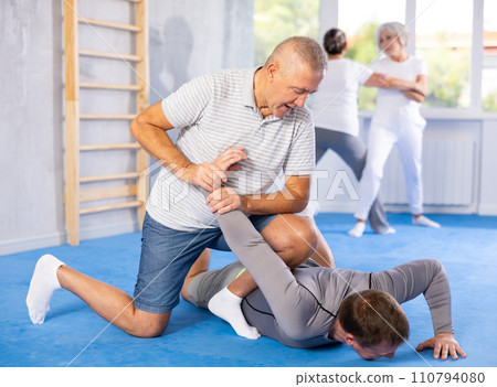 Self-defense lesson - mature man learns how to do painful hold with trainer in gym 110794080