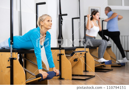 Senior female pensioner training hands and lower back while laying on Pilates Wanda chair in rehabilitation center 110794151