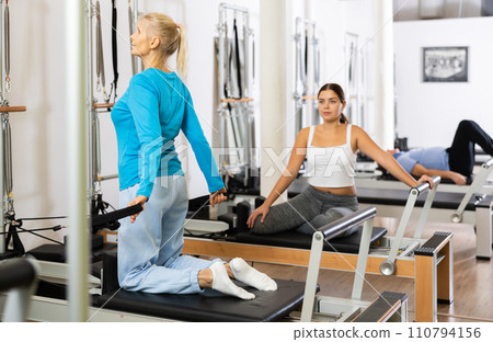 Two sporty focused young mature women in sportswear performing Pilates exercise for back on reformer bed machine in gym Two sporty focused young mature women in sportswear performing Pilates exercise for back on reformer bed machine in gym 110794156