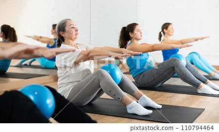 Group of young elderly people in sportswear exercising Pilates with mini balls while lying on mats in rehabilitation center Group of young elderly people in sportswear exercising Pilates with mini balls while lying on mats in rehabilitation center 110794464