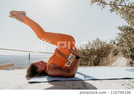 Fitness woman sea. Happy middle aged woman in orange sportswear exercises morning outdoors on yoga mat with laptop in park over ocean beach. Female fitness pilates yoga routine. Healthy lifestyle. Fitness woman sea. Happy middle aged woman in orange sportswear exercises morning outdoors on yoga mat with laptop in park over ocean beach. Female fitness pilates yoga routine. Healthy lifestyle. 110799196