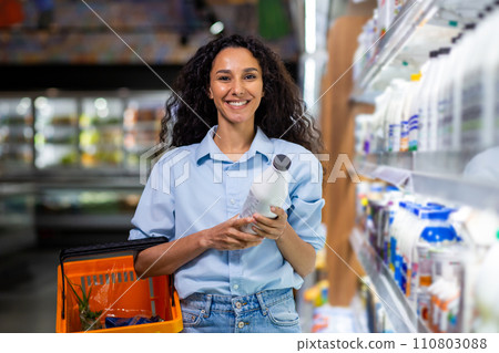 Cheerful customer holding a milk bottle while shopping for groceries, with a basket in a store aisle. Cheerful customer holding a milk bottle while shopping for groceries, with a basket in a store aisle. 110803088
