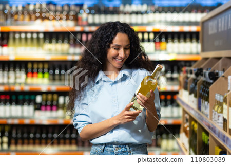 Happy female customer choosing a bottle of wine at a grocery store, examining product quality in the alcohol aisle. Happy female customer choosing a bottle of wine at a grocery store, examining product quality in the alcohol aisle. 110803089