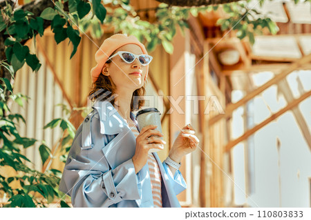 Stylish woman in raincoat and hat with a glass of coffee in hand looks at town district Stylish woman in raincoat and hat with a glass of coffee in hand looks at town district 110803833
