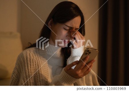A woman whose eyes get tired from looking at her smartphone late at night in a dark room 110803969