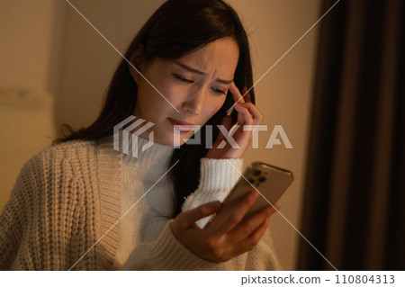 A woman who looks at her smartphone late at night in a dark room gets tired eyes, gets a headache, and is worried about slander. 110804313