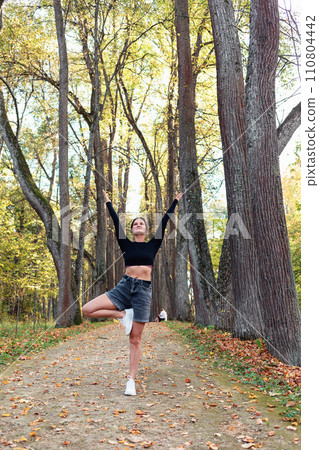 Slender woman performs vrikshasana exercise, tree pose with arms raised and spread to the sides, exercising in casual clothes on a sunny day in the park while standing on a walking path 110804442