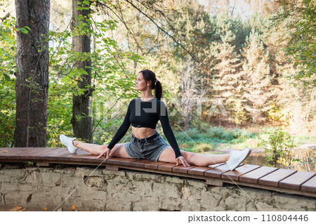 A slender woman performs the exercise Samakonasana, cross split, training in casual clothes on a sunny evening in the park, sitting on the railing of the observation deck A slender woman performs the exercise Samakonasana, cross split, training in casual clothes on a sunny evening in the park, sitting on the railing of the observation deck 110804446