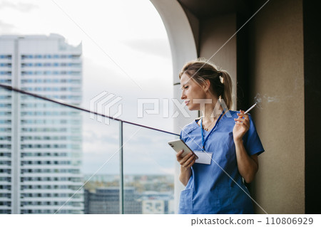 Female nurse smoking cigarette at balcony after work. Doctor feeling overwhelmed at work, taking break. 110806929