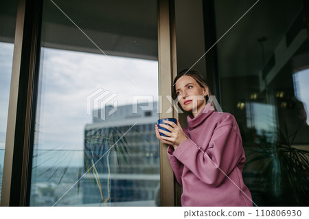 Female nurse enjoying cup of coffee at home after work, standing by window hoodie. Work-life balance for healthcare worker. 110806930