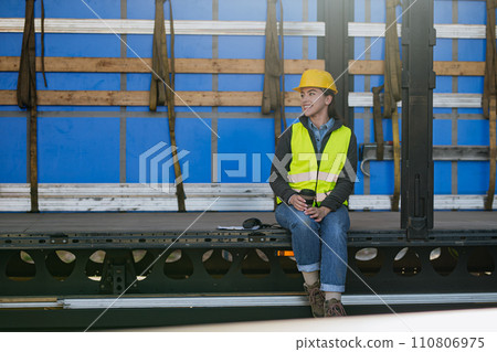 Female warehouse receiver in reflective vest and helmet sitting inside of truck in cargo area, trailer. 110806975