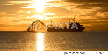 Vessel engaged in dredging at sunset time. Hopper dredger working at sea. Ship excavating material from a water environment. Beautiful sunset. Vessel engaged in dredging at sunset time. Hopper dredger working at sea. Ship excavating material from a water environment. Beautiful sunset. 110807698
