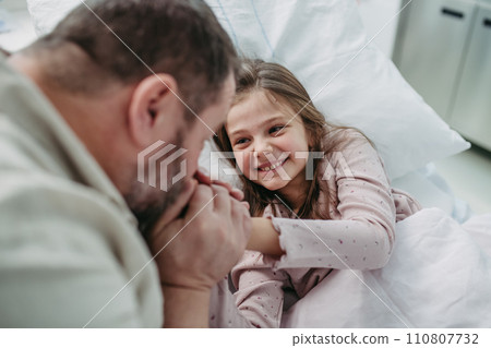 Father sitting beside daughter lying on hospital bed, playing with plush toy. Gift from dad to sick little girl in hospital. 110807732