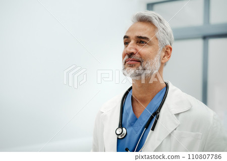 Portrait of confident mature doctor standing in Hospital corridor. Handsome doctor with gray hair wearing white coat, stethoscope around neck standing in modern private clinic, looking at camera. 110807786