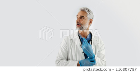 Confident mature doctor putting on medical gloves. Handsome doctor with gray hair wearing white coat, stethoscope around neck. Banner, white background with copy space. 110807787