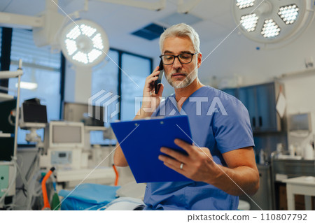 Portrait of confident ER doctor standing in hospital emergency room. Handsome doctor in scrubs holding clipboard, standing in modern private clinic. Portrait of confident ER doctor standing in hospital emergency room. Handsome doctor in scrubs holding clipboard, standing in modern private clinic. 110807792