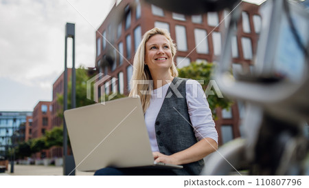 Businesswoman, freelancer or manager working outdoors in city park. Woman with laptop on knees making call on smartphone, sitting on park bench during sunny day. Concept of working remotely. 110807796