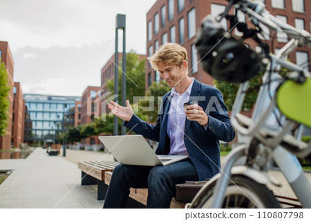 Businessman, freelancer or manager working outdoors in city park. Man with laptop on knees drinking coffee, having video call, listening music. Concept of working remotely. Businessman, freelancer or manager working outdoors in city park. Man with laptop on knees drinking coffee, having video call, listening music. Concept of working remotely. 110807798