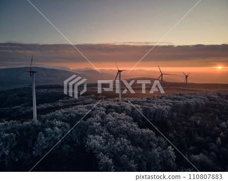 Aerial landscape photography of sunrise over frost-covered nature with wind turbines. Windmill in soft morning light with icy trees around. Concept of wind power as clean, renewable energy source. 110807883