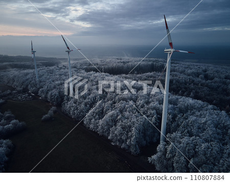 Aerial landscape photography of sunrise over frost-covered nature with wind turbines. Windmill in soft morning light with icy trees around. Concept of wind power as clean, renewable energy source. 110807884