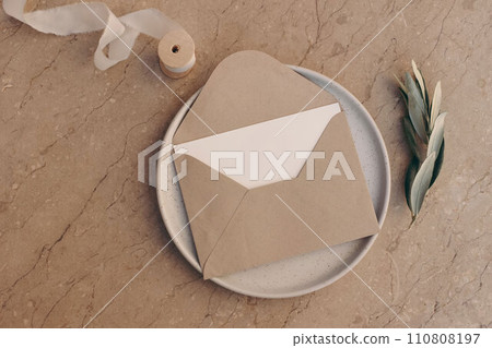Mediterranean wedding stationery. Birthday mock-up. Blank greeting card, envelope on ceramic plate. Olive tree branch, silk ribbon. Beige marble background, floor. Summer holiday design. Flatlay, top 110808197